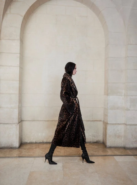 Woman in a long animal-print coat and black boots walks in a hallway with arched openings, side view.