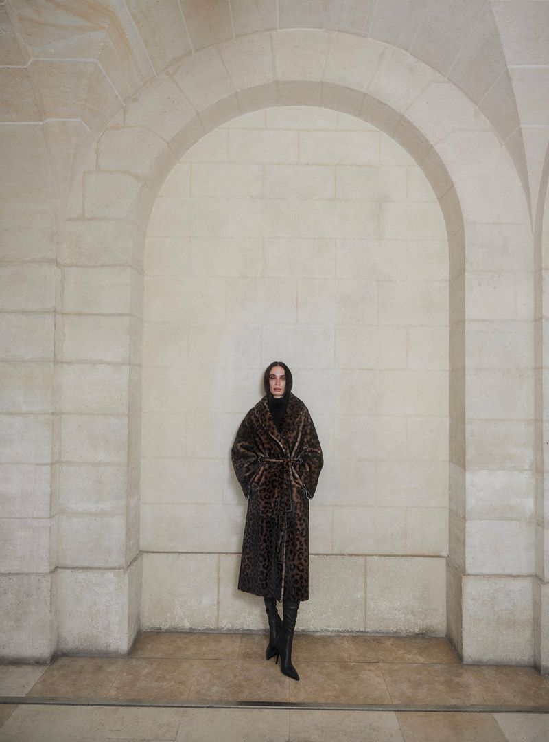 Model standing centered inside a tall stone archway wearing a leopard print coat and dark boots