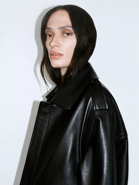 Portrait of a woman with dark hair in a black leather trench coat, posing against a white wall in a photo studio setting with natural lighting.