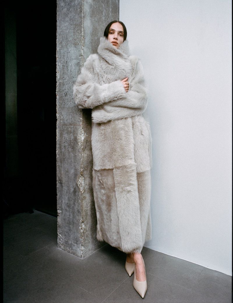 Elegant adult woman poses indoors wearing a full-length, oversized grey shearling coat with matching pumps against a modern wall.
