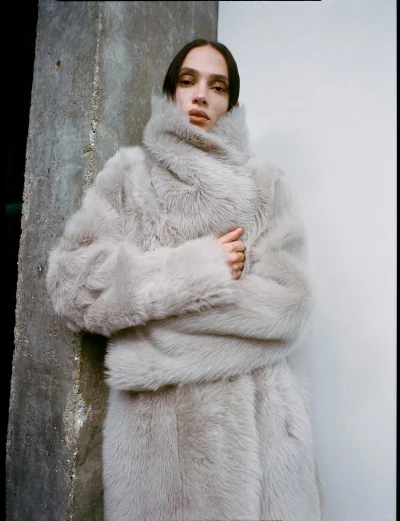 Model wearing a plush light grey faux fur coat with a high collar, standing against a concrete pillar and white wall.