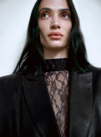 Close-up portrait of a female model with dark hair wearing a black leather blazer over a lace top against a plain light background