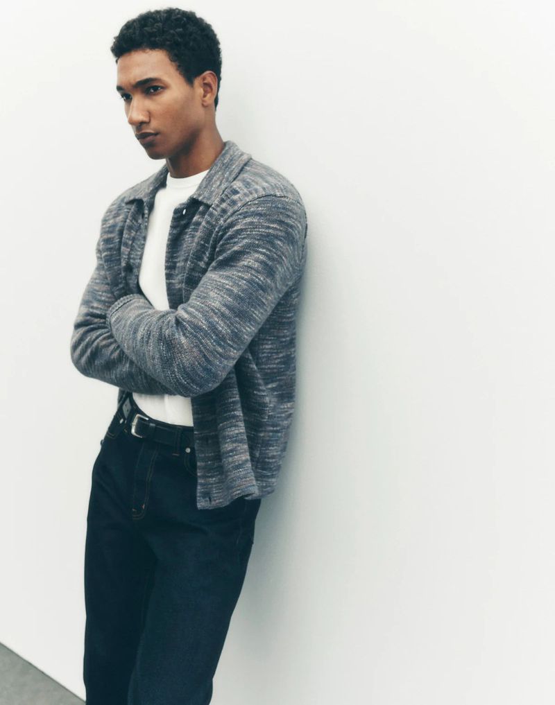 Adult male with crossed arms leans against a white wall wearing a collared heather grey shirt and black pants in a medium studio shot.