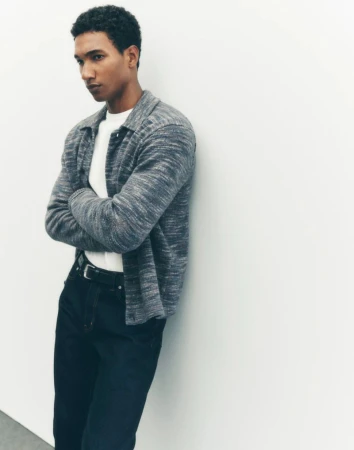 Adult male with crossed arms leans against a white wall wearing a collared heather grey shirt and black pants in a medium studio shot.
