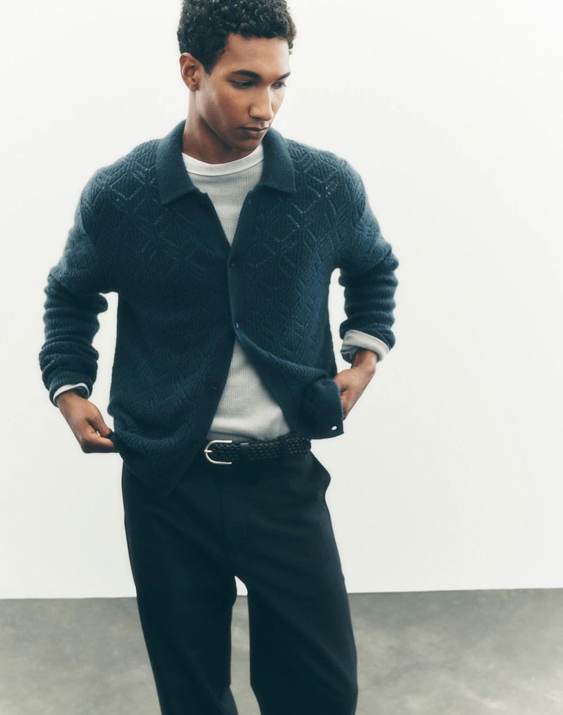 Young man wearing a patterned cardigan and black pants in a studio setting, posing and looking to the side.