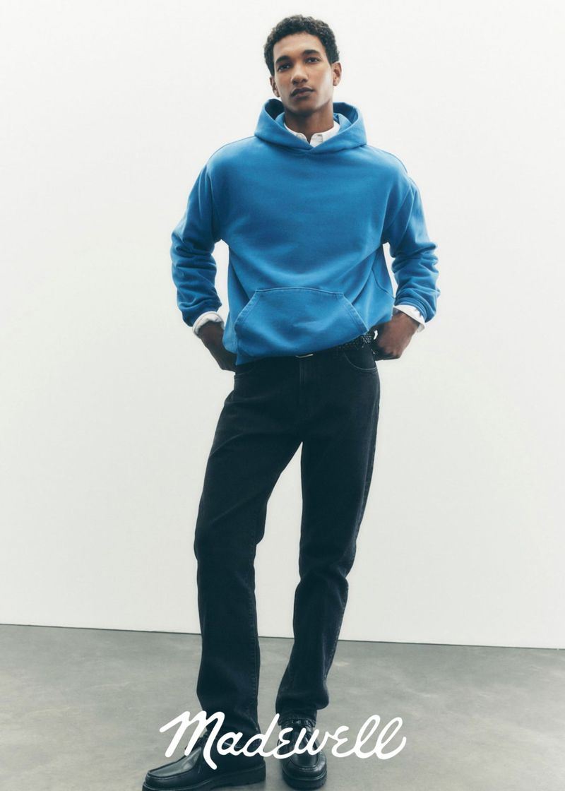 A young adult male poses in a blue hooded sweatshirt, dark jeans, and black shoes against a white backdrop in a studio setting.