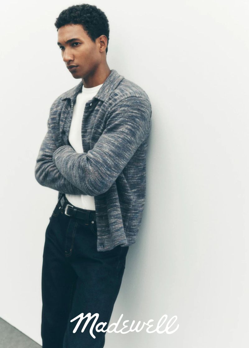 A young adult male poses against a white wall, wearing a gray shirt jacket over a white tee, and dark jeans, showcasing a casual fashion aesthetic.
