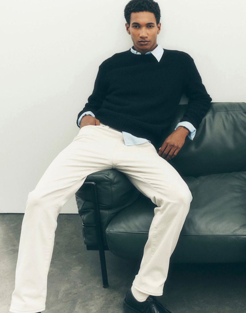 Male model wearing black sweater and white pants seated on a dark leather sofa against a white wall.