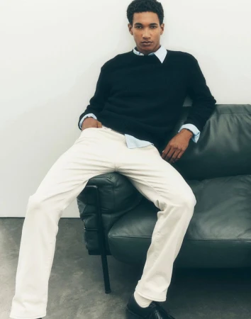 Male model wearing black sweater and white pants seated on a dark leather sofa against a white wall.