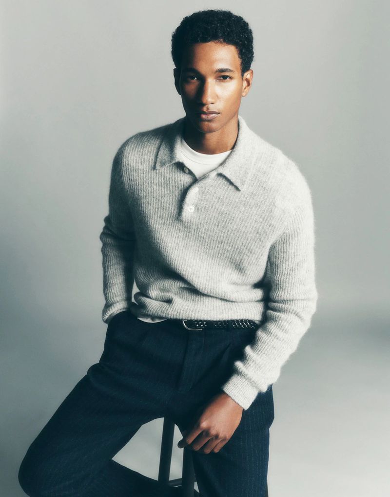A young adult male poses indoors wearing a grey wool sweater and dark trousers with a belt, sitting against a neutral background.