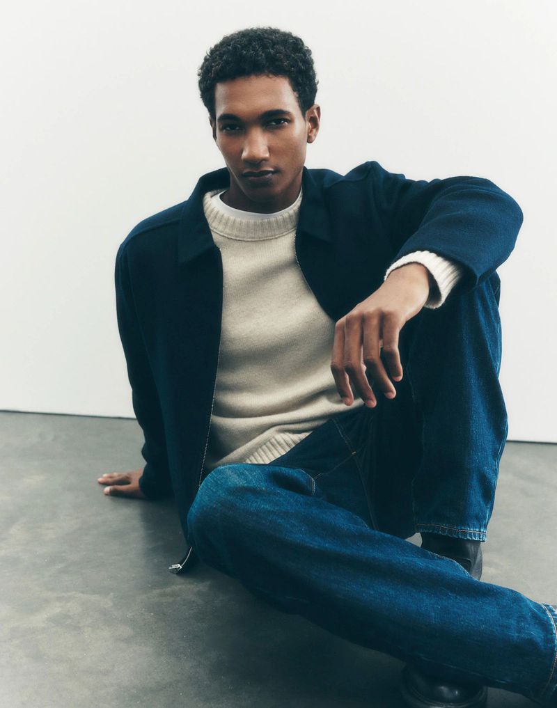 A casually dressed Black male model with an open navy blue jacket sits on a gray surface looking at the camera in a studio shot.