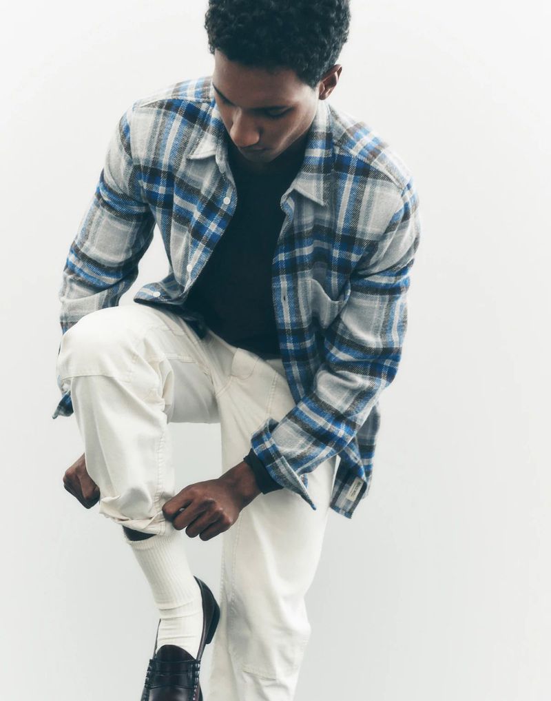 An adult male adjusts his cream-colored pants, sitting against a bright white backdrop, wearing a flannel shirt and leather loafers.