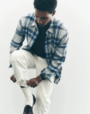 An adult male adjusts his cream-colored pants, sitting against a bright white backdrop, wearing a flannel shirt and leather loafers.