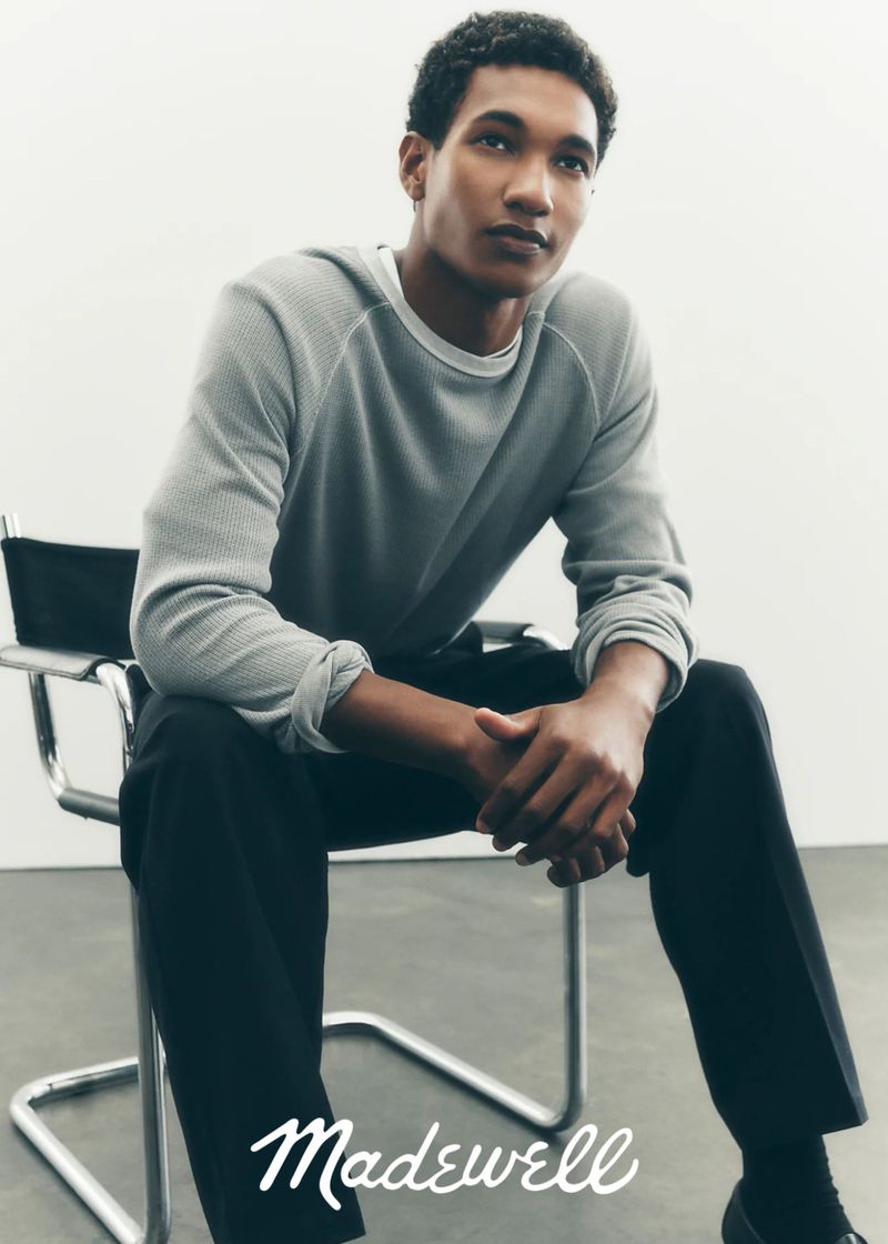 Studio shot of a young adult Black man wearing a grey long sleeve shirt and black pants, sitting on a chair, looking off camera.