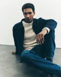 Man wearing a dark jacket, cream knit sweater, and blue jeans seated against a white background