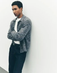 Man with arms crossed wearing a grey knit shirt and dark jeans against a white background
