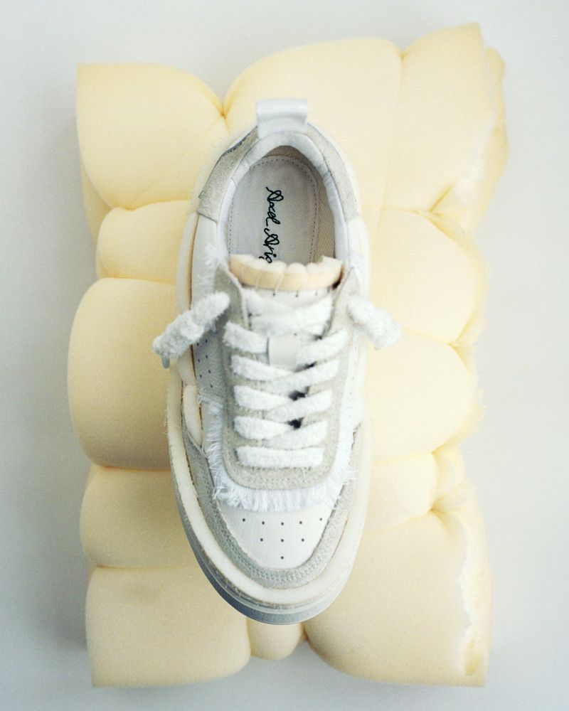 Fashion shot of a single white sneaker with laces on a light yellow puffy pillow against a white background for product marketing.