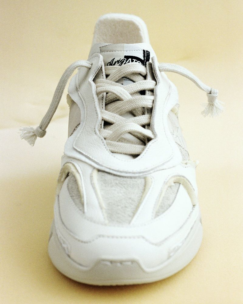 Close-up product shot of a white leather and fabric sneaker with white laces, on a pale yellow background in a studio setting.