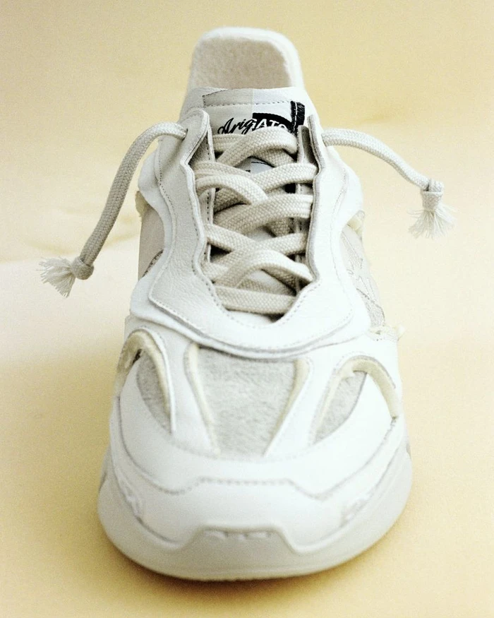 Close-up front view of a white Axel Arigato sneaker with textured panels and loose knotted laces on a plain background