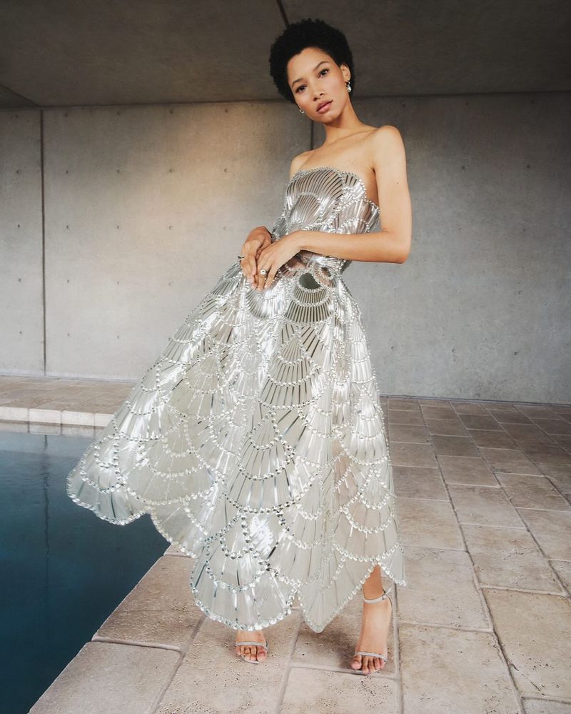 Adult female in a silver strapless gown poses on a patio next to a pool, creating a glamorous fashion shot with concrete backdrop.