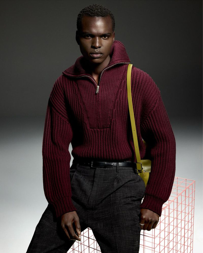 Fashion portrait of a Black man wearing a maroon sweater, grey pants, and a yellow purse, posing against a grey background with a wireframe prop.