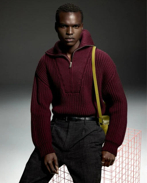 Fashion portrait of a Black man wearing a maroon sweater, grey pants, and a yellow purse, posing against a grey background with a wireframe prop.