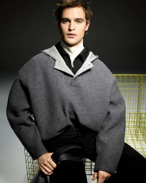 Portrait of a fashionable adult male wearing a grey hooded poncho and black pants, posed in front of a grey background in a studio.