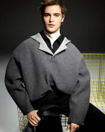 Portrait of a fashionable adult male wearing a grey hooded poncho and black pants, posed in front of a grey background in a studio.
