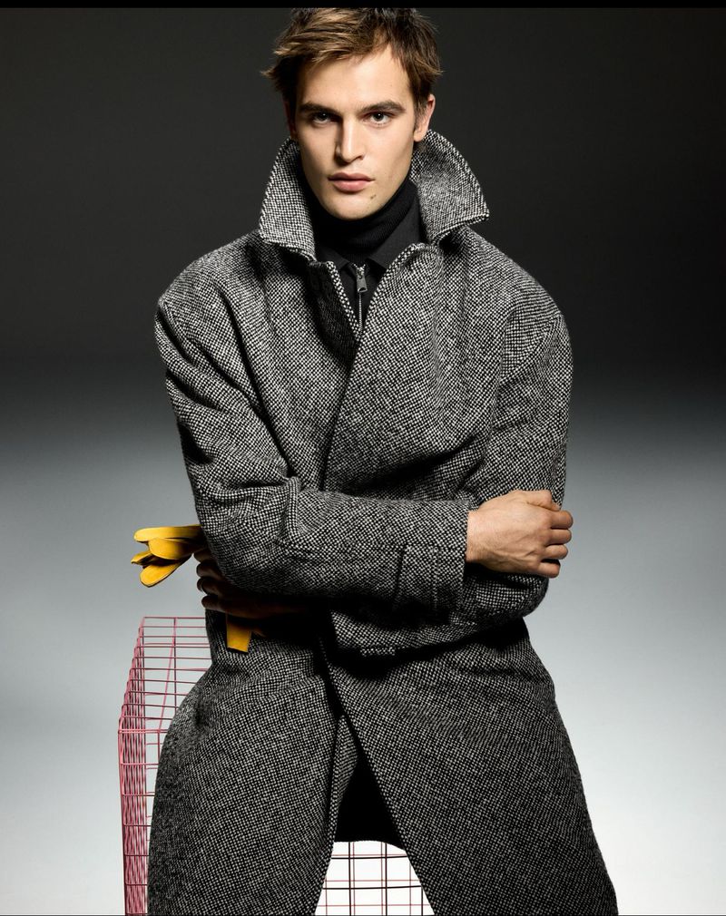 Fashion shot of a man wearing a grey wool coat and black turtleneck, posing next to a wire pet cage in a photo studio with grey backdrop.