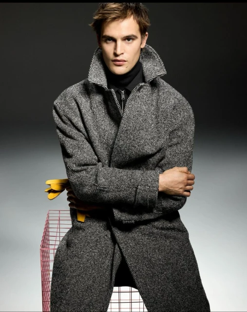 Fashion shot of a man wearing a grey wool coat and black turtleneck, posing next to a wire pet cage in a photo studio with grey backdrop.