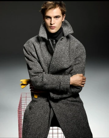 Fashion shot of a man wearing a grey wool coat and black turtleneck, posing next to a wire pet cage in a photo studio with grey backdrop.