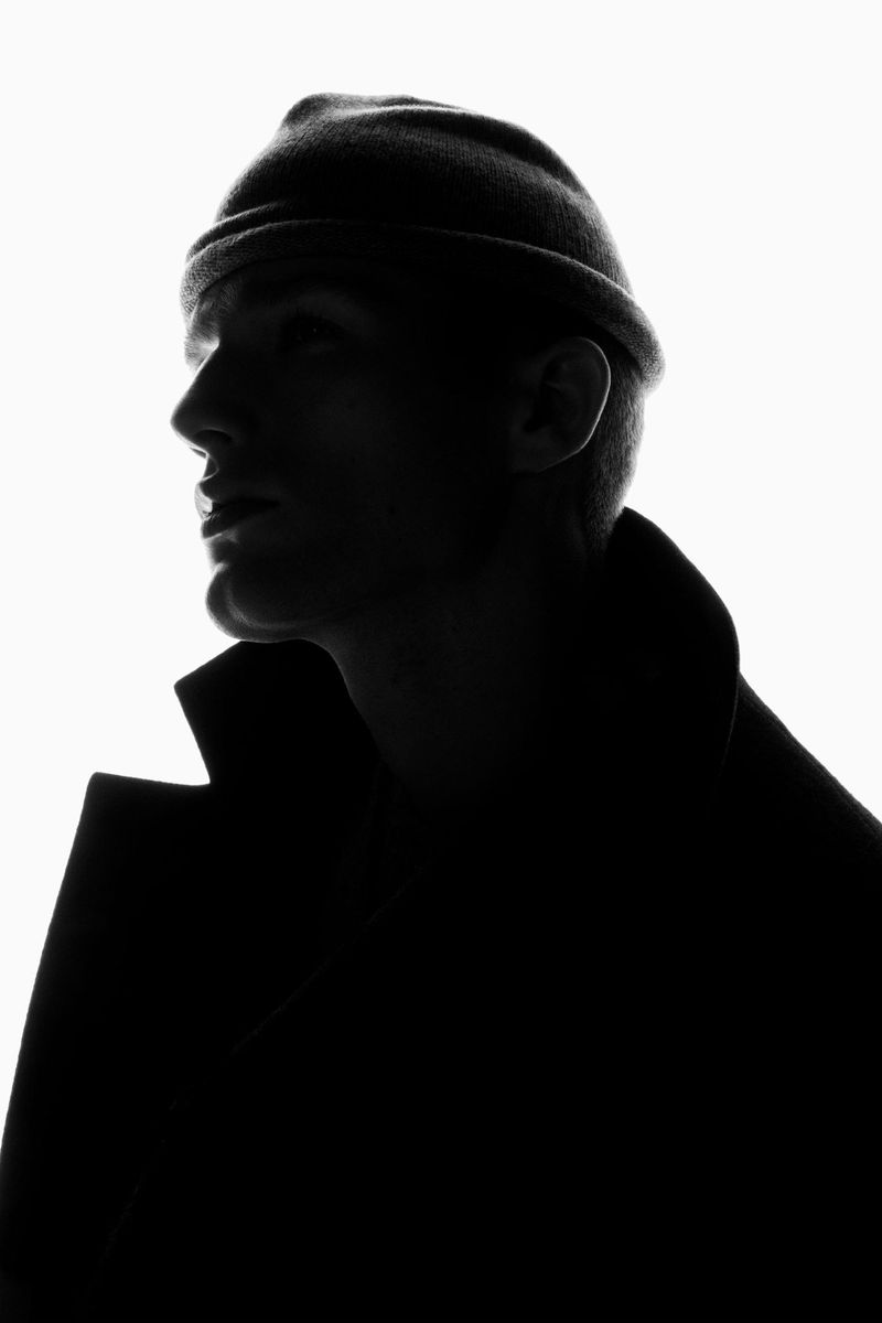 Monochrome portrait of a silhouetted man wearing a beanie and coat against a bright white background in a studio setting with side lighting.