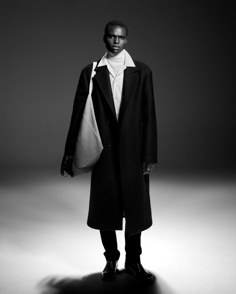 Black and white portrait of a Black male model in a stylish overcoat holding a white tote bag, posed in a studio with neutral backdrop.