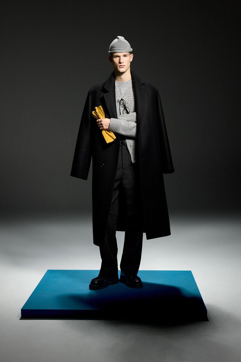 Male fashion model poses in black overcoat and beanie against a gray background, holding yellow gloves, standing on a blue platform.