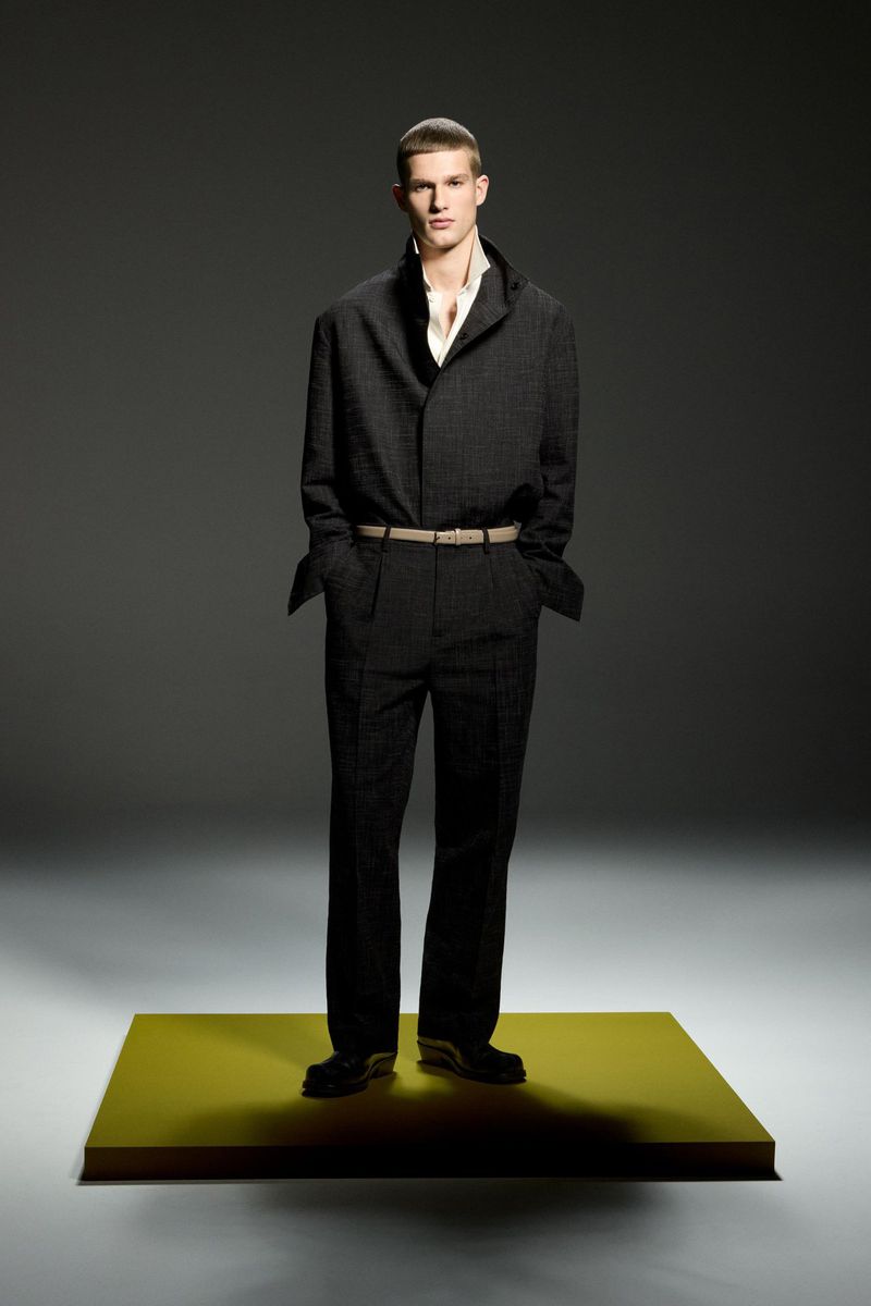Male fashion model poses in dark suit with tan belt and dress shoes on yellow platform in studio with grey backdrop. Full shot.