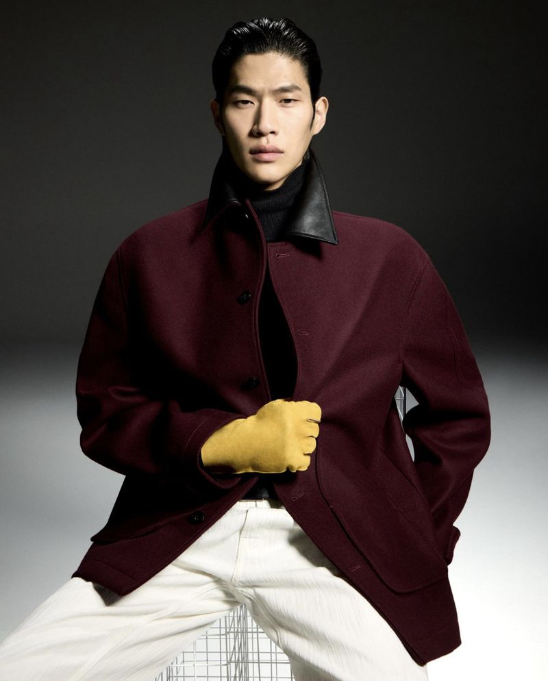 Portrait of a handsome Asian male model in a maroon jacket and white pants, posing in a studio with moody lighting for a fashion shoot.