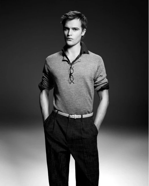 Black and white portrait of a male model in a polo shirt and dark slacks, hands in his pockets, eyeglasses hanging on his shirt.