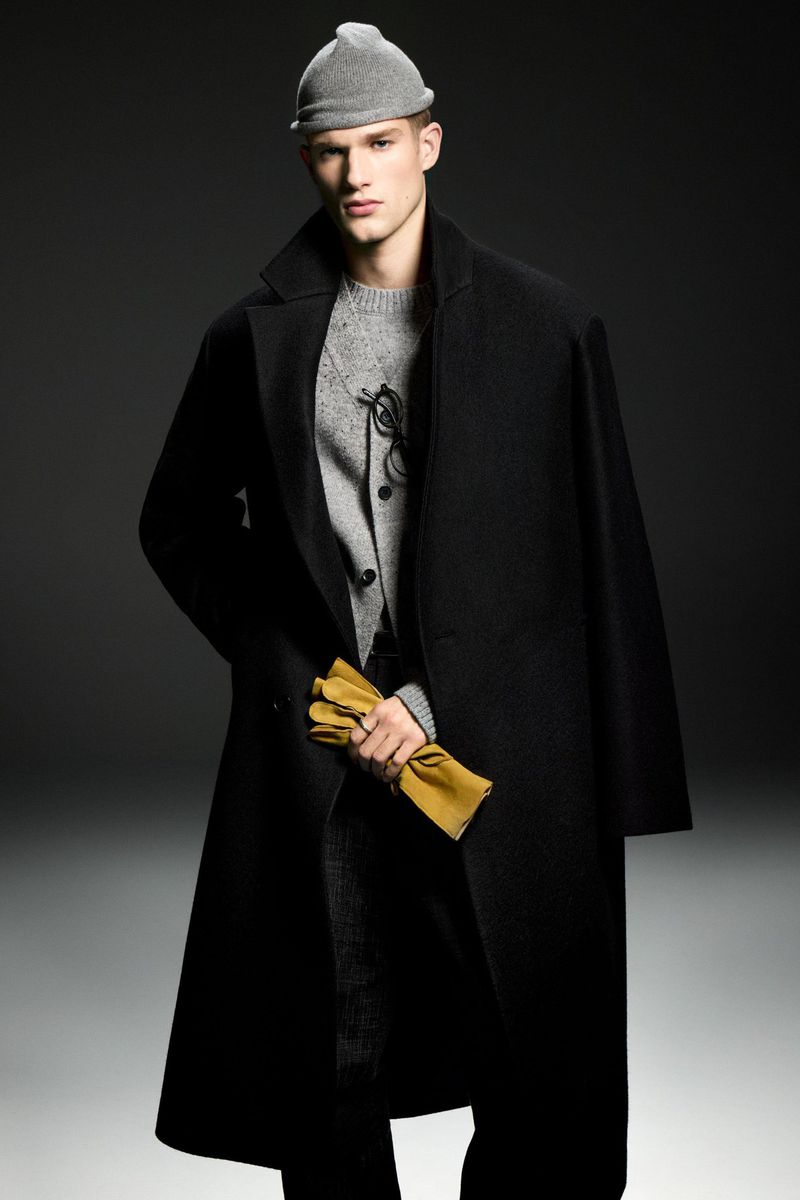 Fashionable adult man in overcoat, beanie, and gloves posing indoors against dark background, shot at eye level in medium framing.