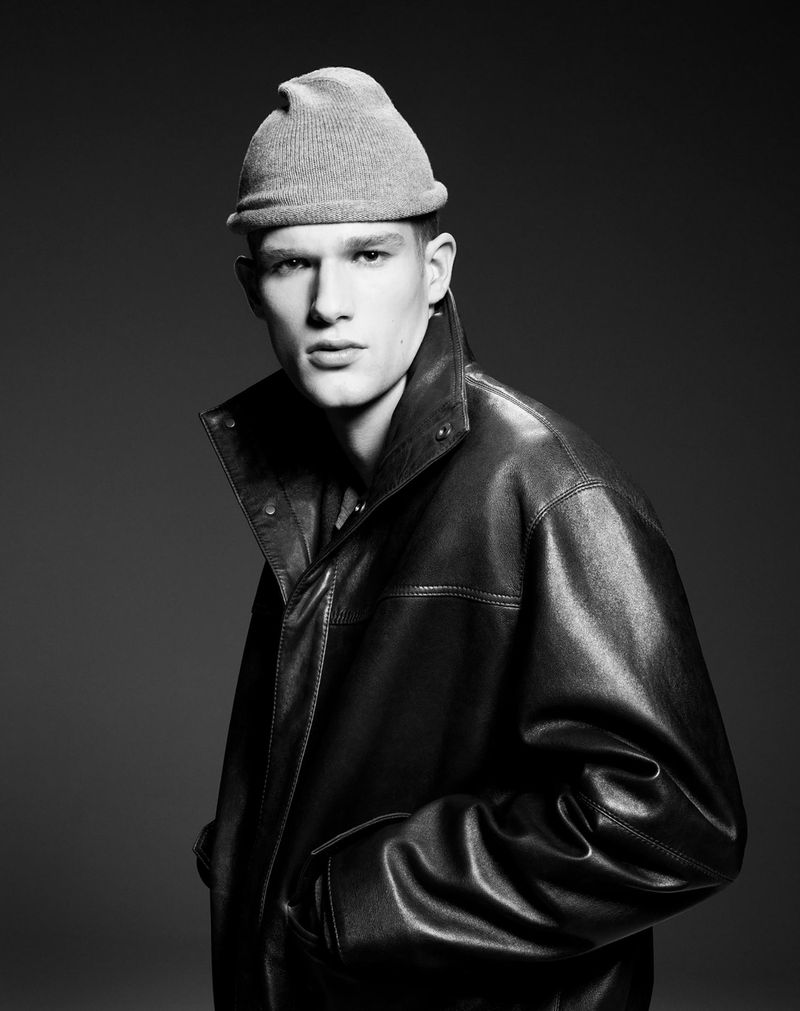 Black and white portrait of a young adult male wearing a beanie and a leather jacket, standing indoors with a neutral expression.