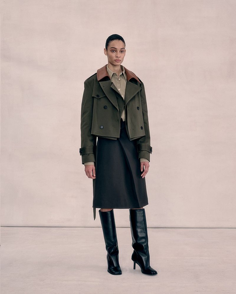 A woman in an olive trench coat and dark skirt stands in a full shot in a photo studio with off-white walls and bright, neutral lighting.