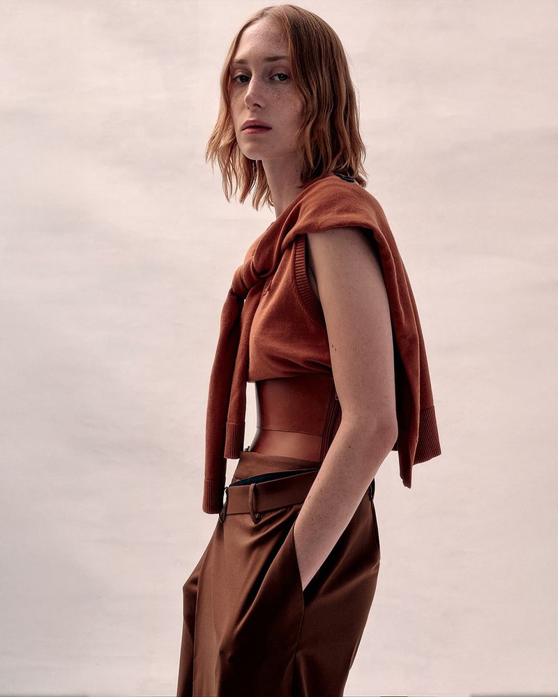 Fashionable woman poses in copper sleeveless crop top and matching high-waisted trousers with a light sweater draped over her shoulders.