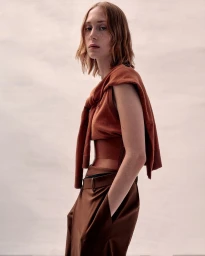 Model with shoulder-length reddish hair wearing a rust-colored sleeveless top and brown trousers against a neutral background.