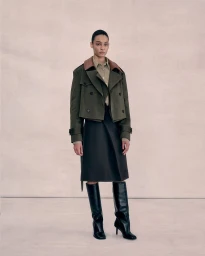 Model wearing an olive green cropped jacket and black skirt standing against a neutral studio backdrop