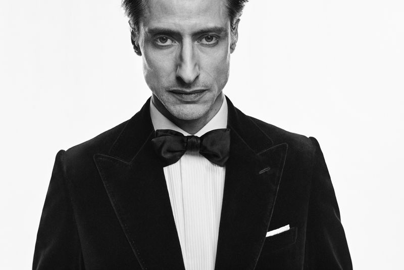 Black and white portrait of a man in a tuxedo with a bow tie, close-up shot in a brightly lit photo studio against a white backdrop.