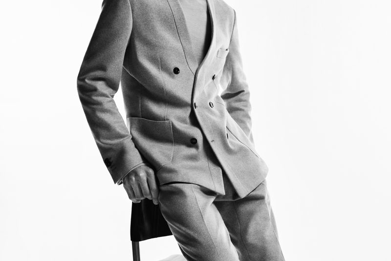 Monochrome shot of a person leaning against a chair, wearing a grey double-breasted suit against a bright white background in a studio.