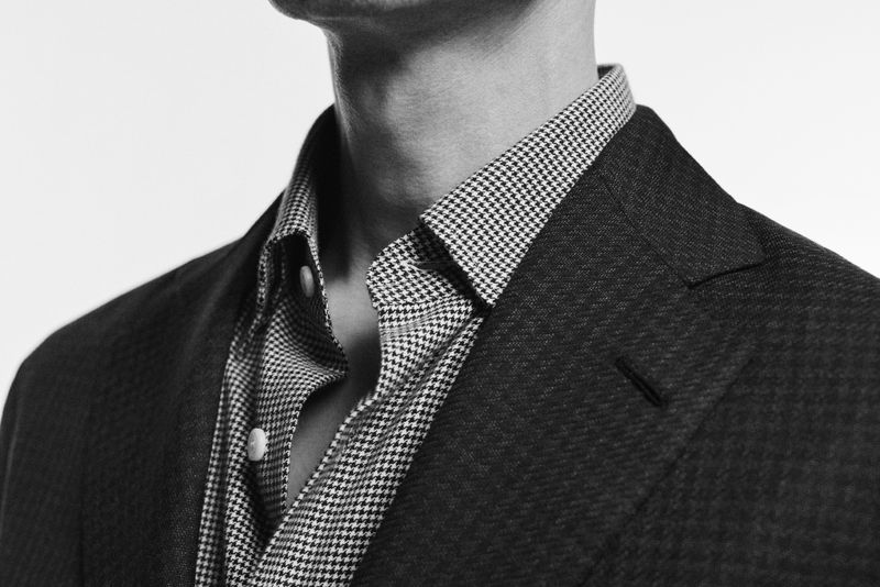 Torso of an adult male wearing a houndstooth patterned shirt and a dark colored suit jacket against a white background.