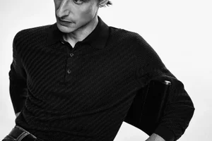 Close-up portrait of a man wearing a black textured knit polo shirt against a plain white background for Brioni FW 24.