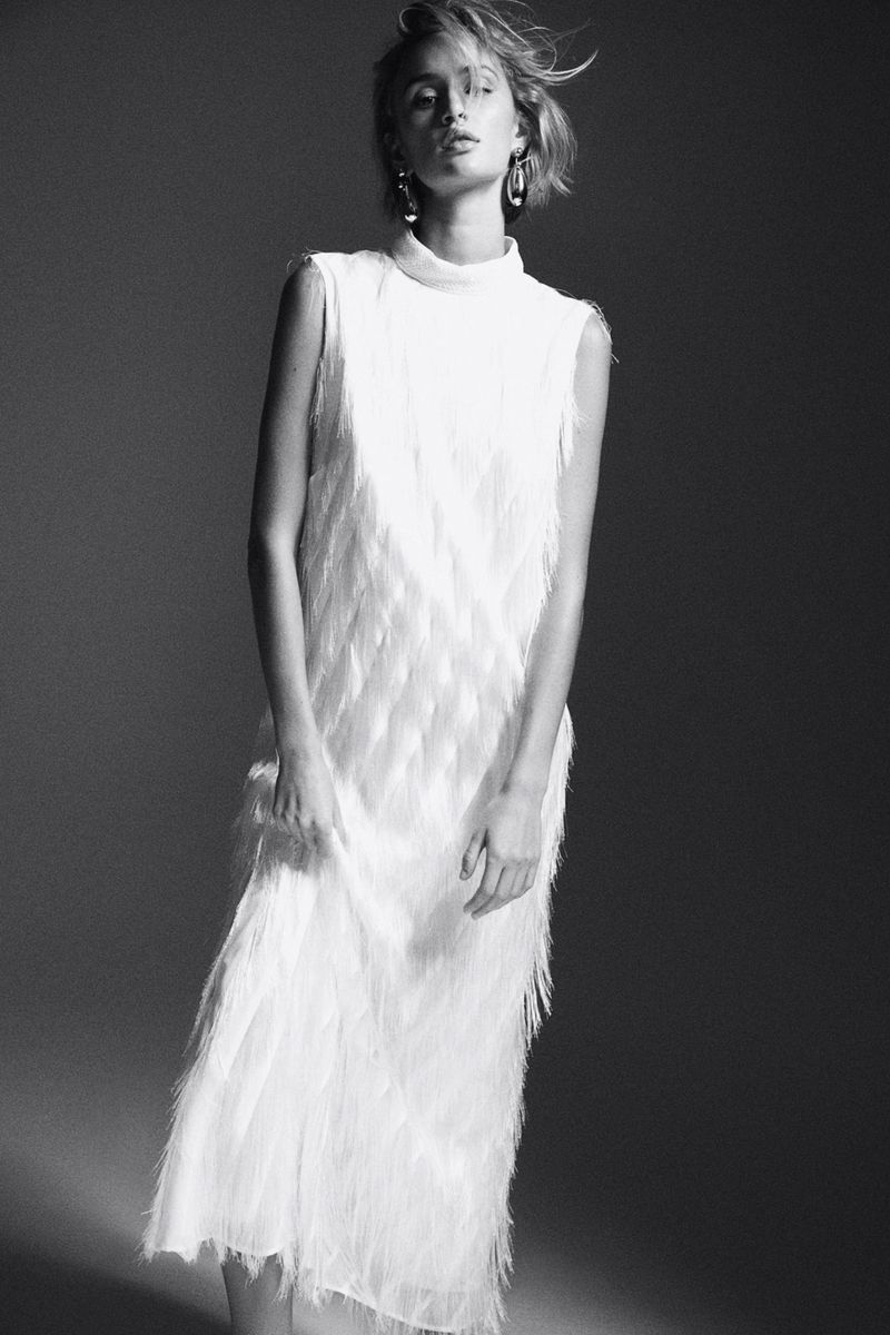 Model wearing a white fringed sleeveless dress with statement earrings in a monochrome studio lookbook image