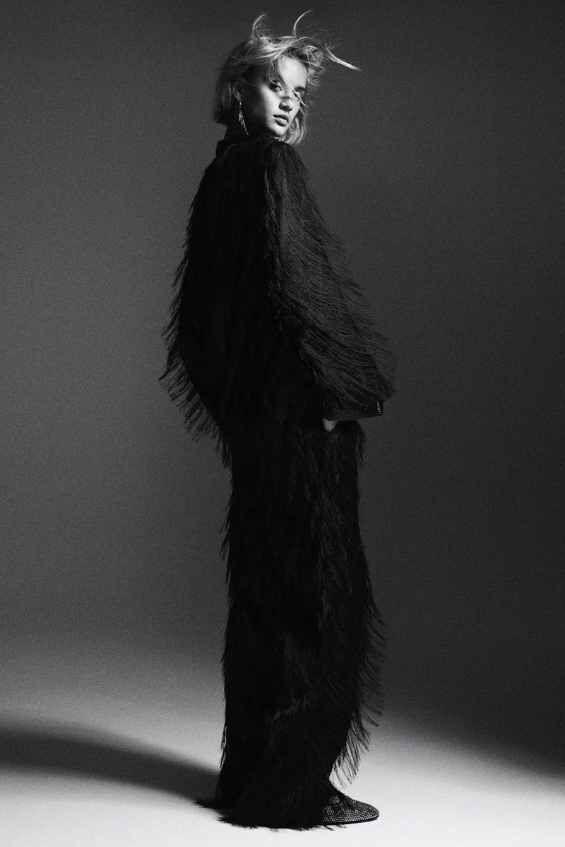 Monochrome studio shot of a stylish young adult female in a black fringe dress, posing in a three-quarter view against a gray background.