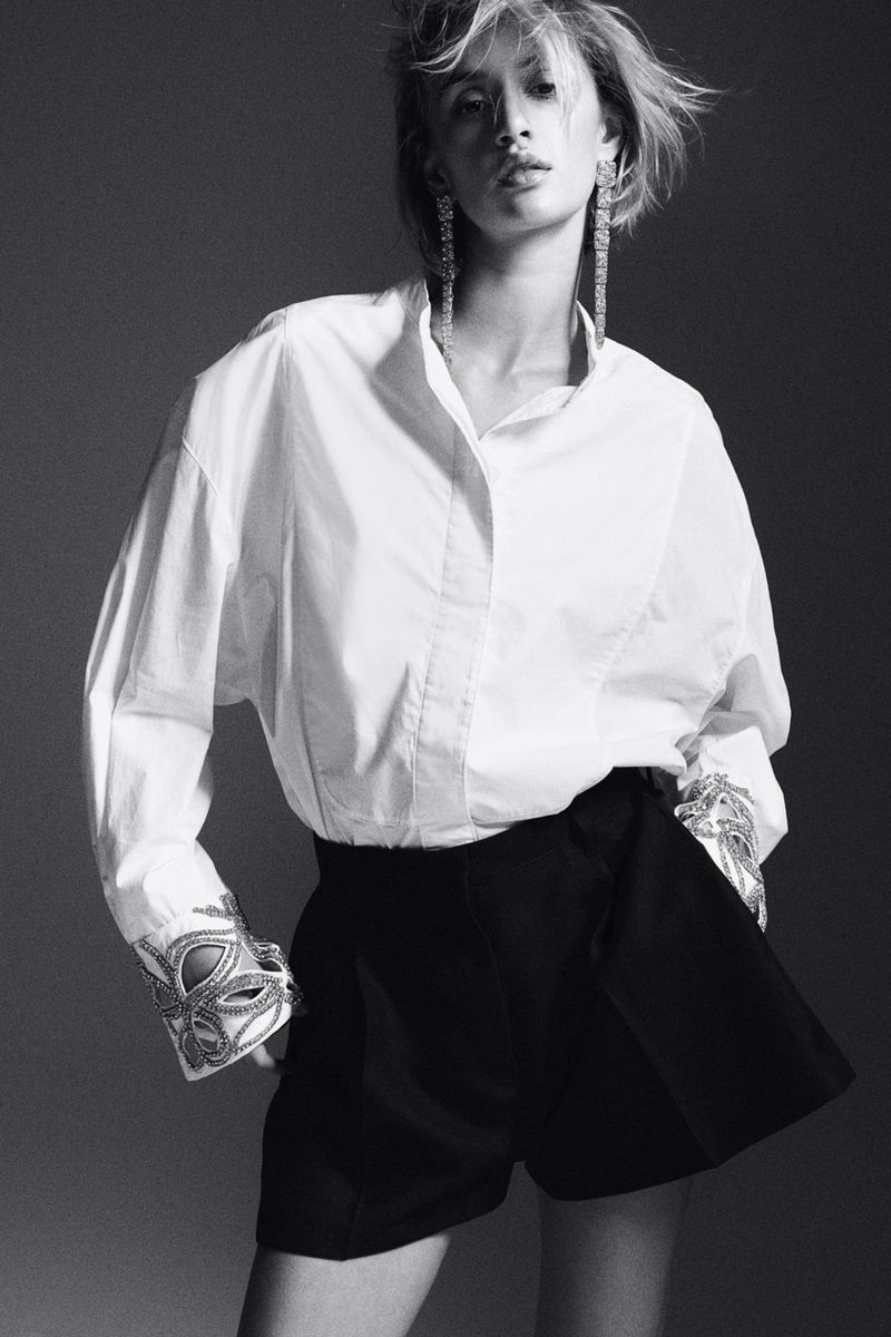 Fashion shot of a woman wearing a white shirt and black shorts, accessorized with long earrings and decorative cuffs in a grey studio.